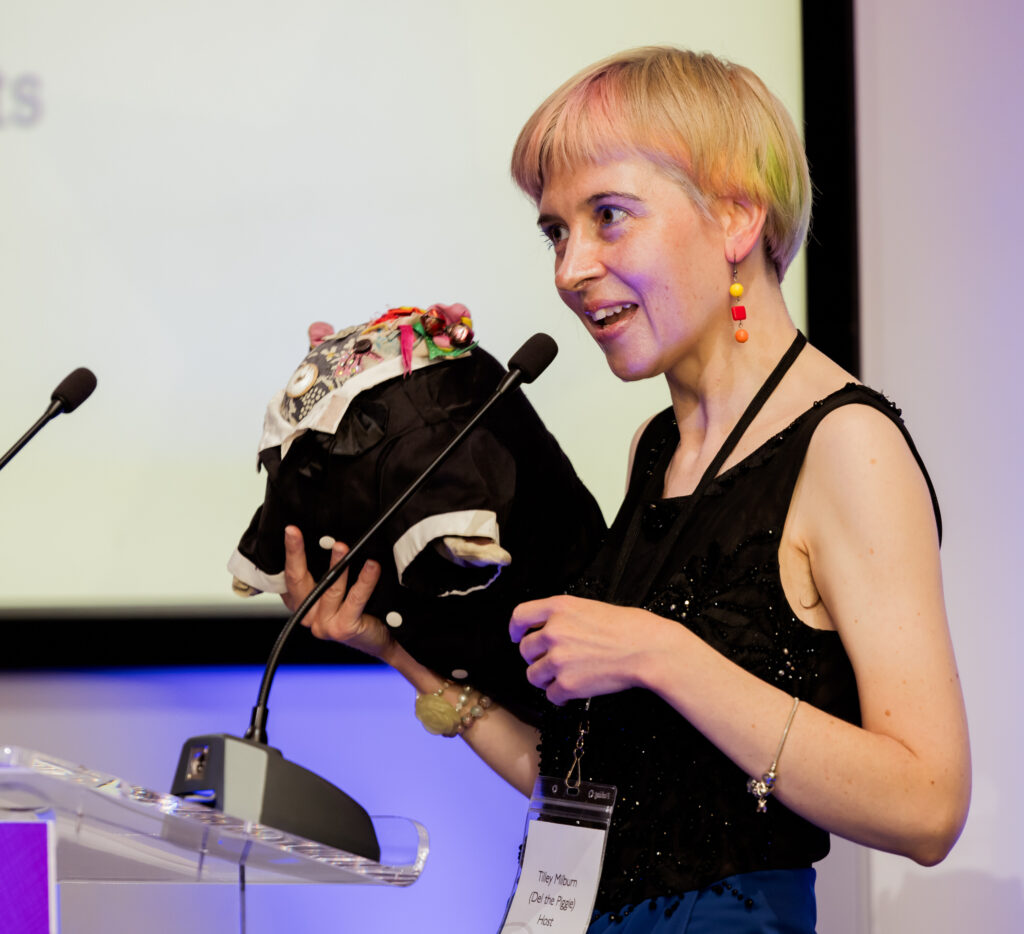 A woman with short blonde hair wearing a smart black dress on a stage holding a toy pig wearing a tuxedo