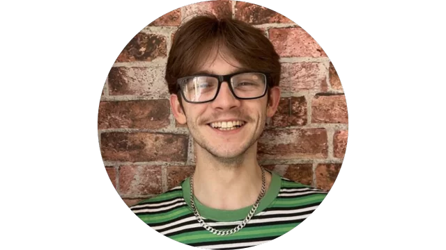 Man smiling with brown hair and glasses standing in front of a brick wall. He is wearing a green and black striped shirt and a silver chain necklace.